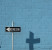 A 'One Way' sign creates a shadow in the shape of a cross against a cinder block wall that has been painted blue.